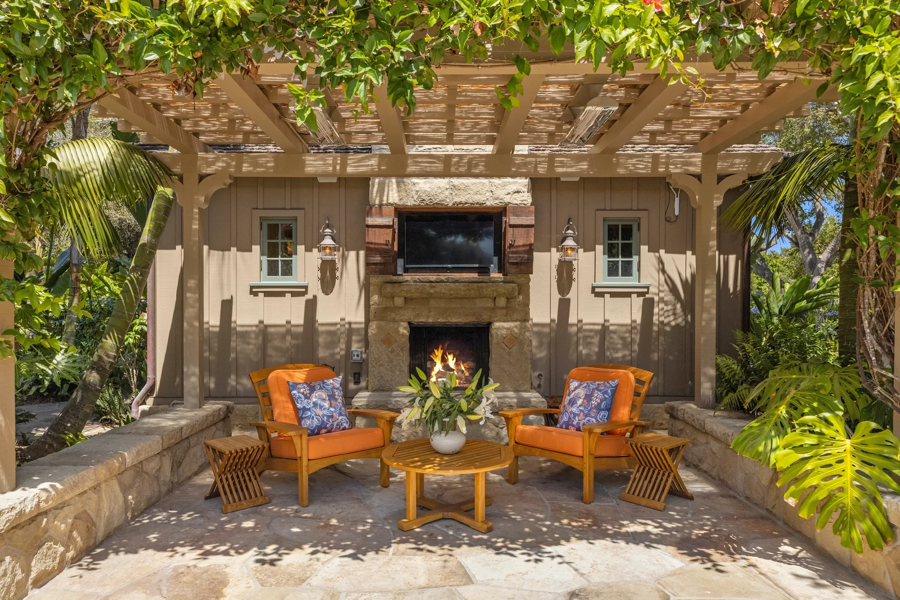 An outdoor seating area beneath a wooden pergola, with a fireplace and tropical plants.