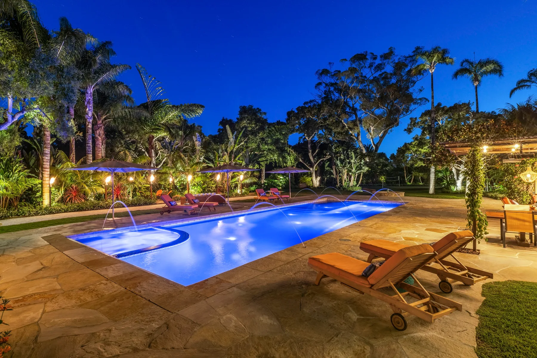 A swimming pool is surrounded by lounge seating and tropical landscaping at night.