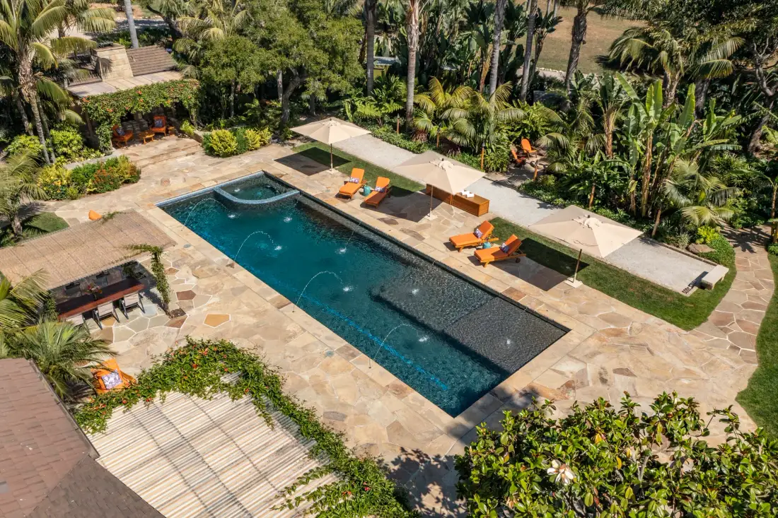 An outdoor swimming pool sits among paved terraces, lounge seating, and tropical greenery.