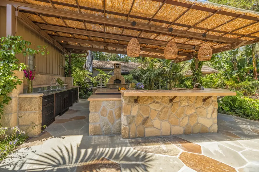 A covered outdoor kitchen with a stone island and surrounding greenery.