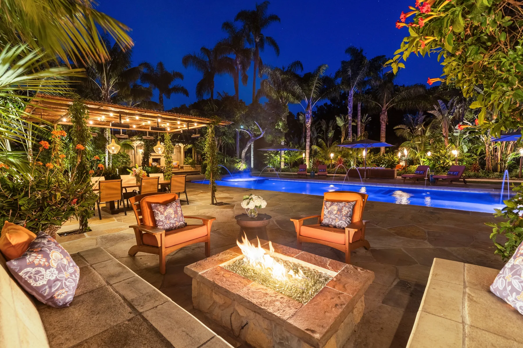 An illuminated outdoor poolside patio with seating and a glowing fire pit.