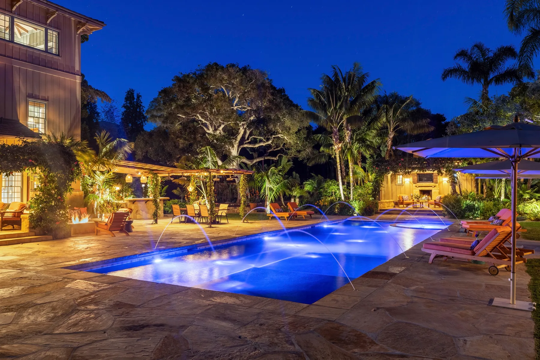 A swimming pool glows at night beside a palm-lined outdoor seating area.