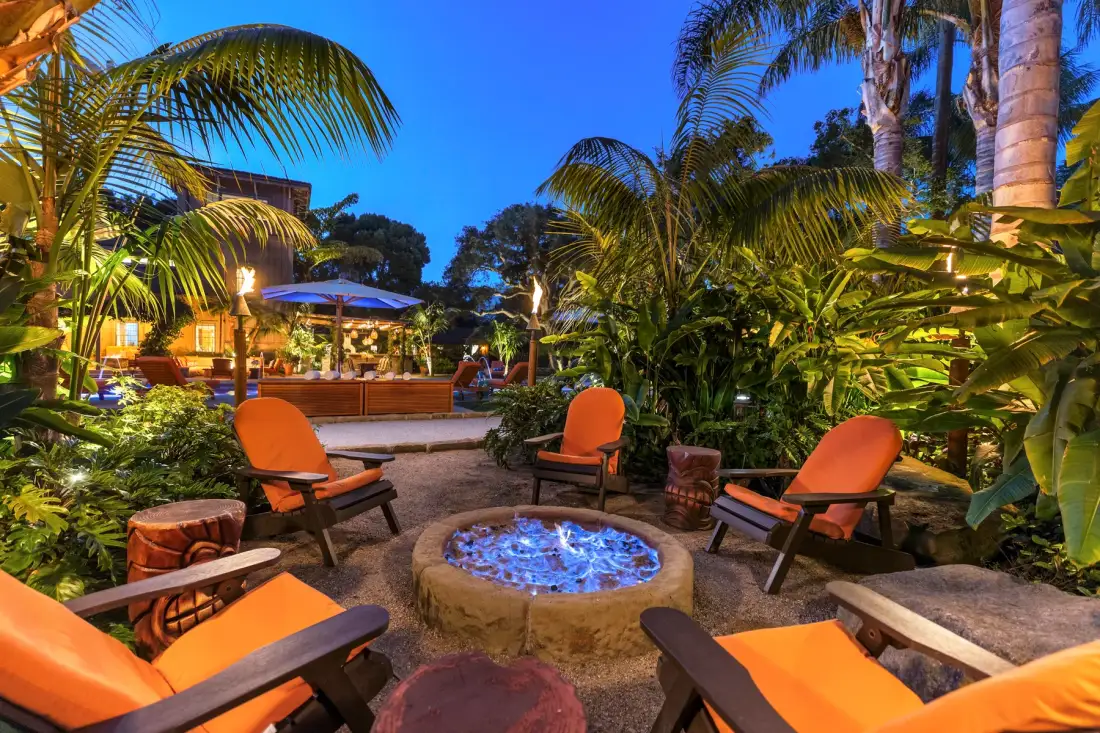 An outdoor garden seating area with orange chairs arranged around a glowing fire pit.