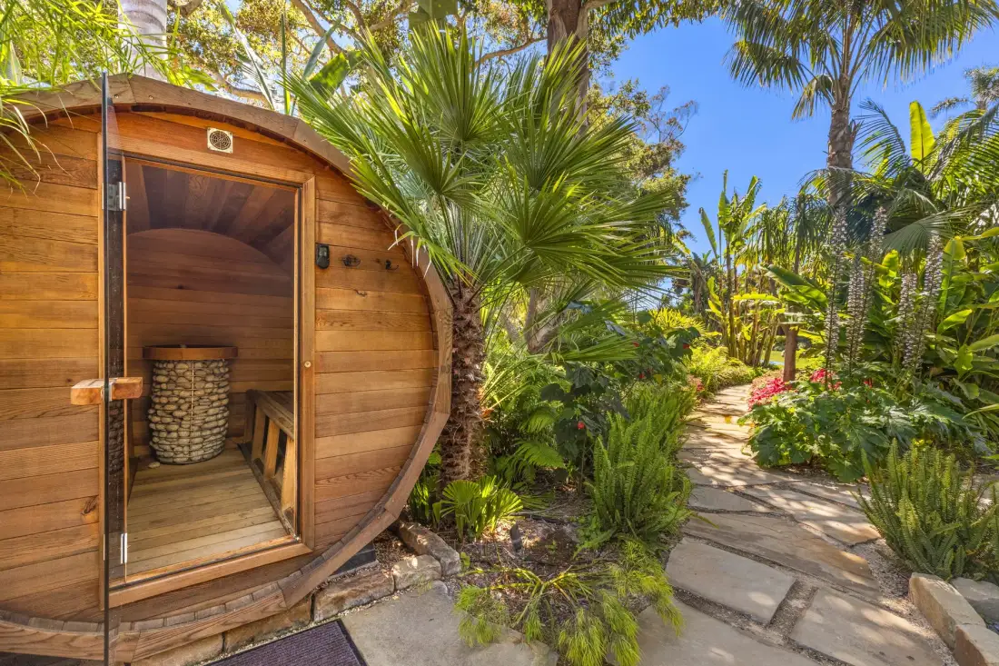 A wooden sauna sits beside a garden path surrounded by tropical plants.