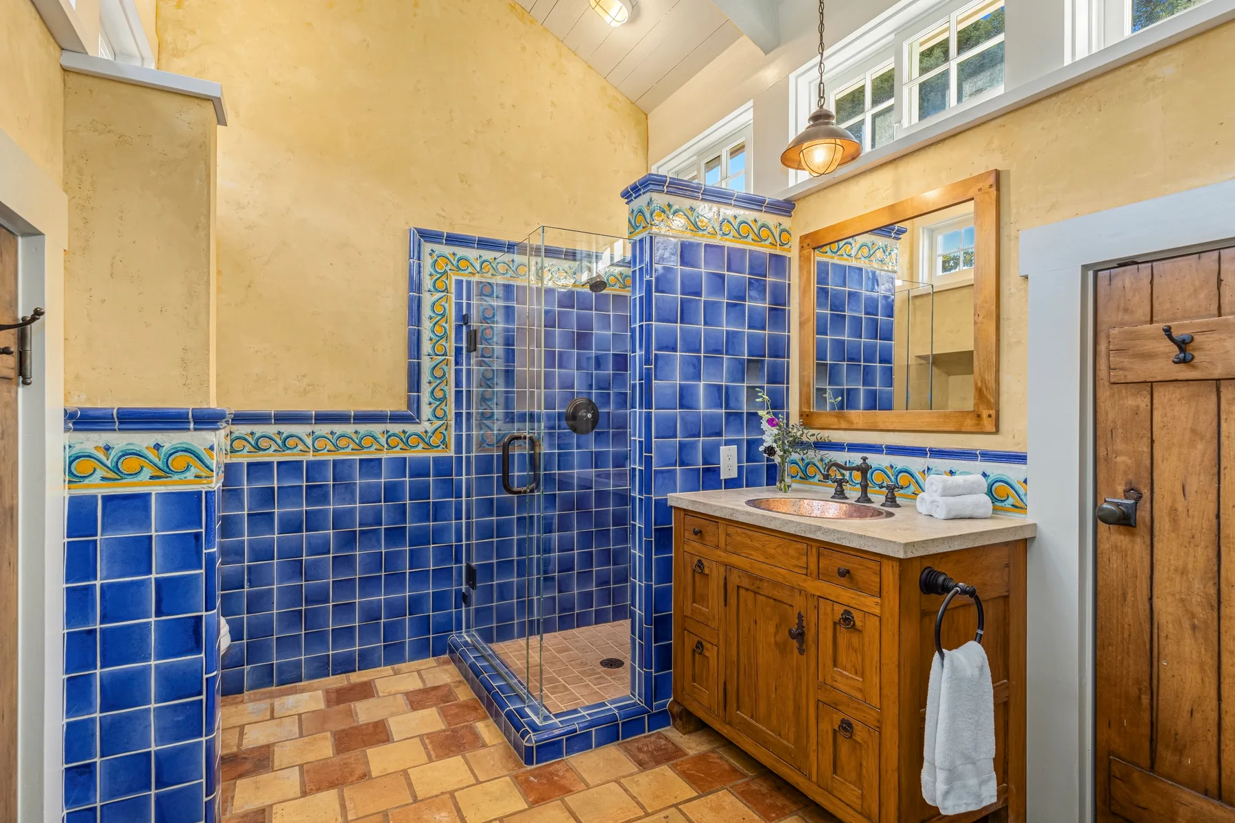 A bathroom with a blue-tiled shower, wooden vanity, and mirror.