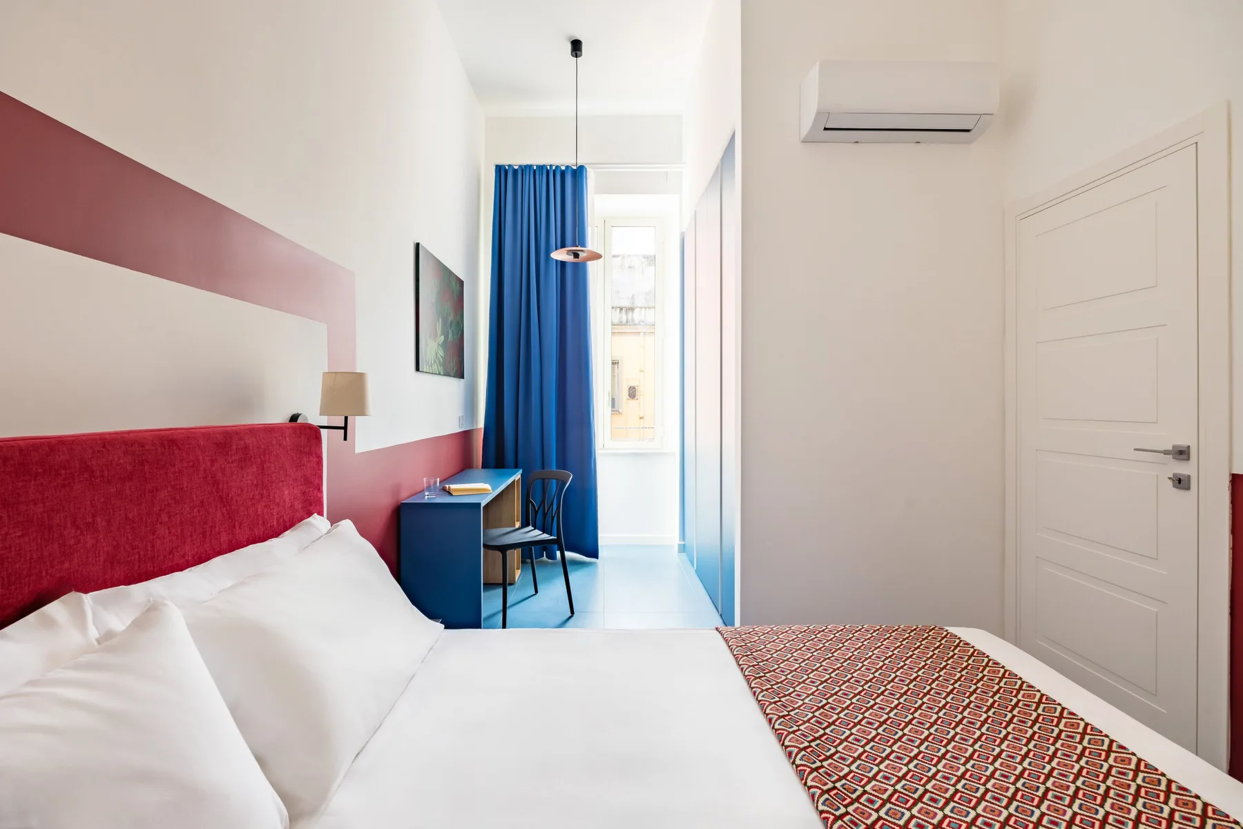 A bedroom with a bed, blue curtains, and a small desk by the window.