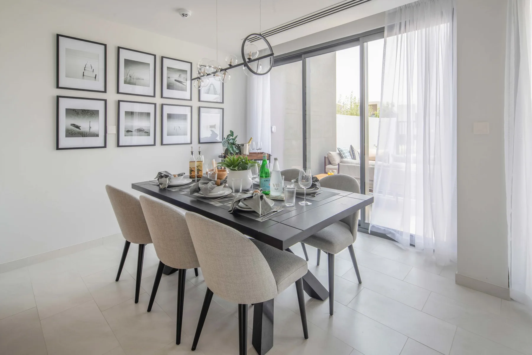 Un comedor tiene la mesa puesta junto a grandes ventanas con cortinas translúcidas.