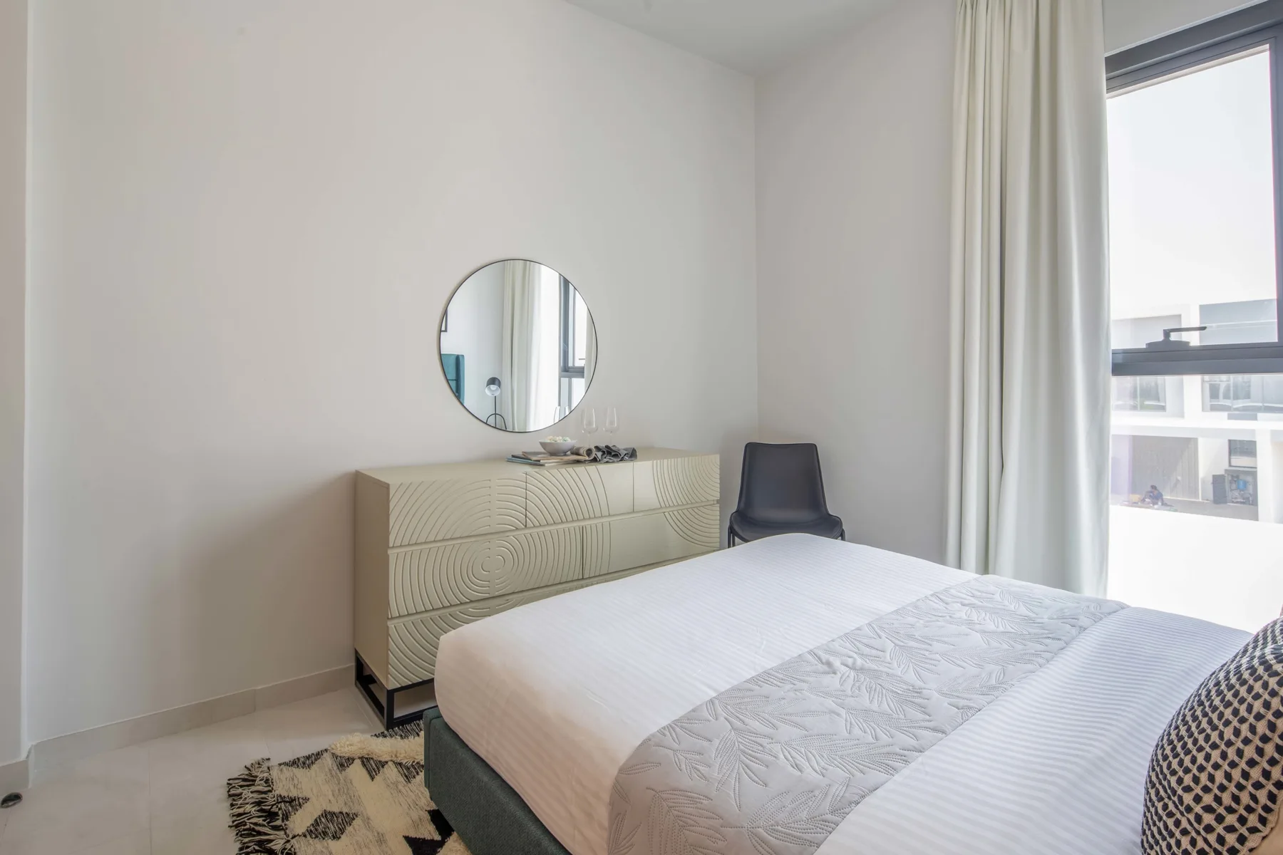 Un dormitorio luminoso con una cama tendida, una cómoda y una ventana grande.