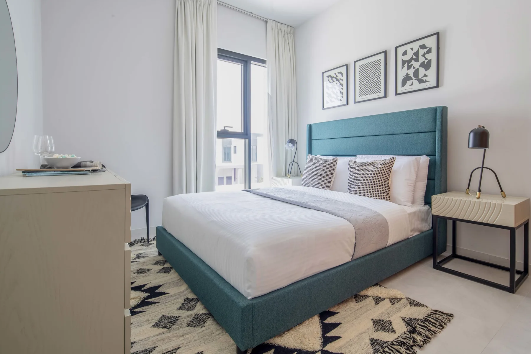 Un dormitorio luminoso con una cama tapizada en verde azulado y una alfombra estampada.