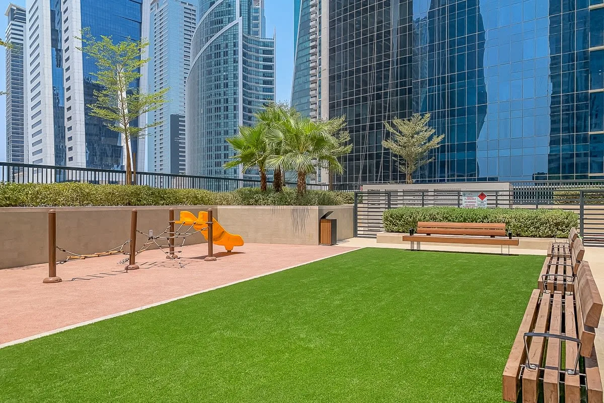 Una zona recreativa al aire libre cuenta con césped verde, bancos y juegos naranjas.