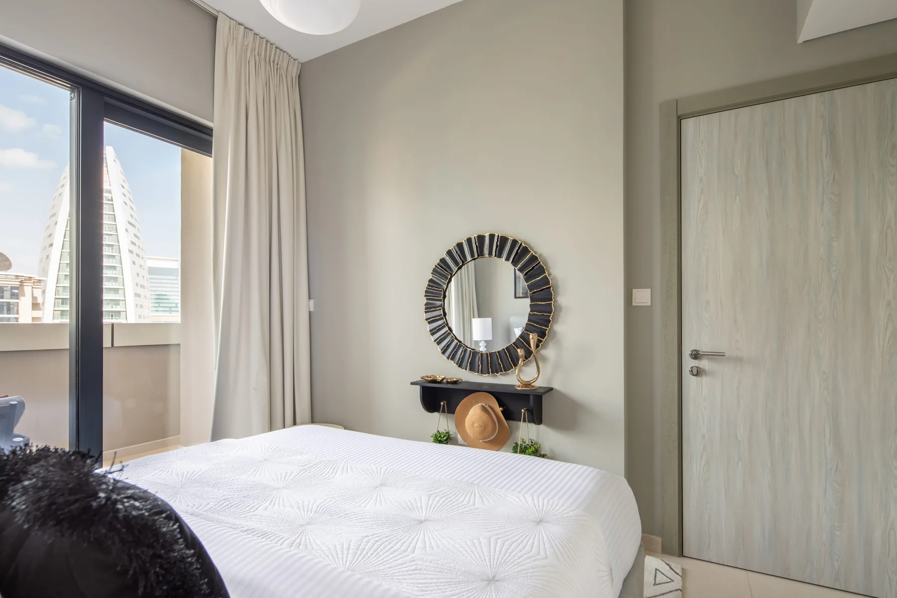 Un dormitorio con una cama blanca, un espejo de pared y una ventana con cortinas.