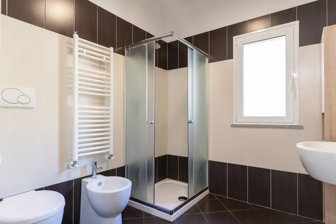Un baño con azulejos, una ducha de vidrio, una ventana y sanitarios blancos.