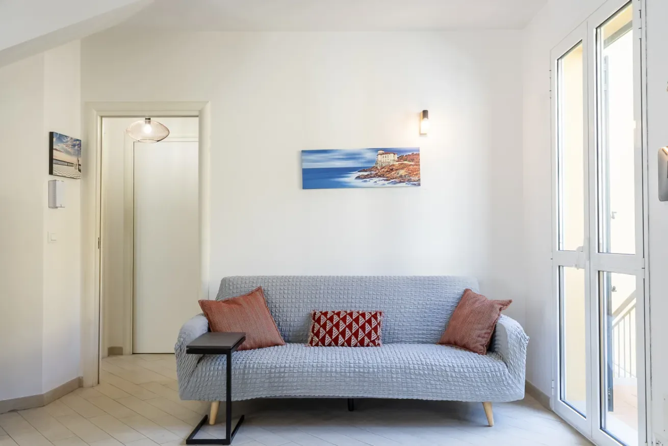 Una sala luminosa con un sofá bajo un cuadro costero y ventanas altas.
