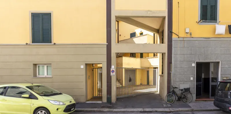 La entrada de un edificio amarillo da a un patio, con coches y bicicletas cerca.