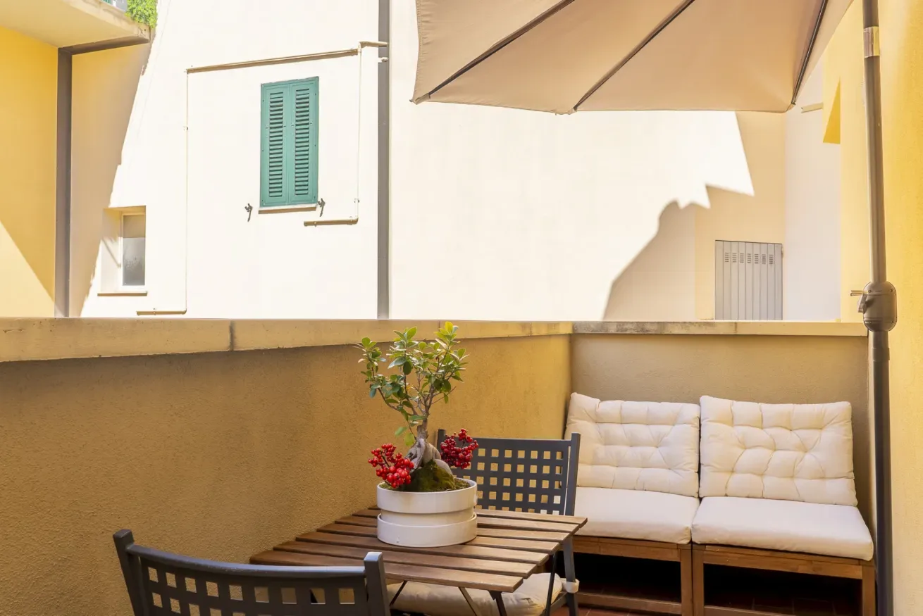 Una zona de estar en un patio al aire libre tiene una mesa de madera, un banco con cojines y una sombrilla.