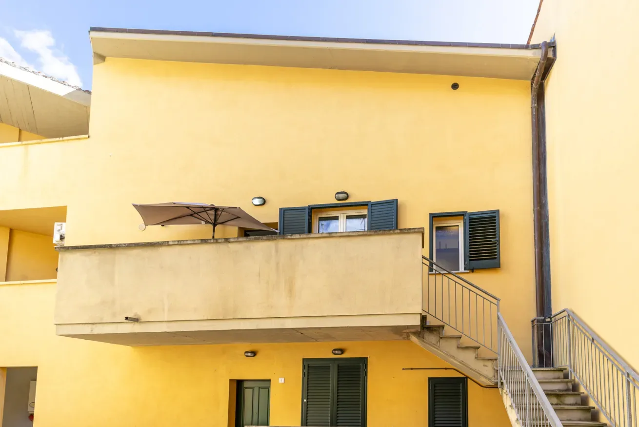 Fachada de un edificio amarillo con balcón, sombrilla y escaleras exteriores.