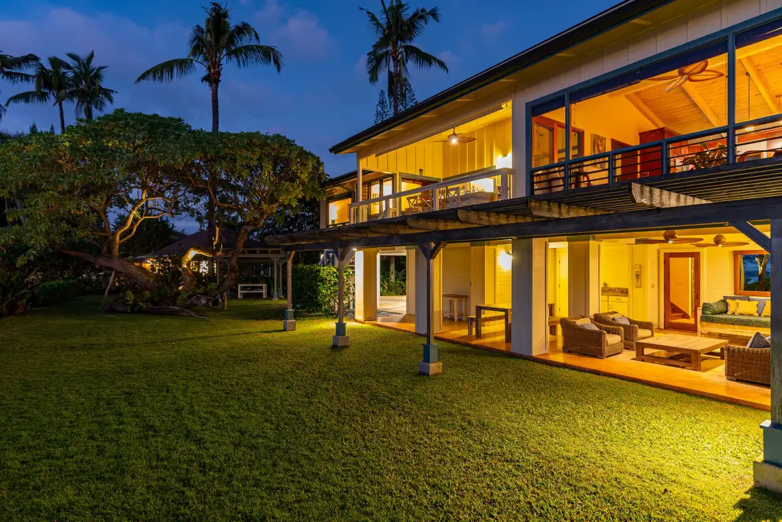 Una casa moderna iluminada al anochecer, junto a un césped y palmeras.