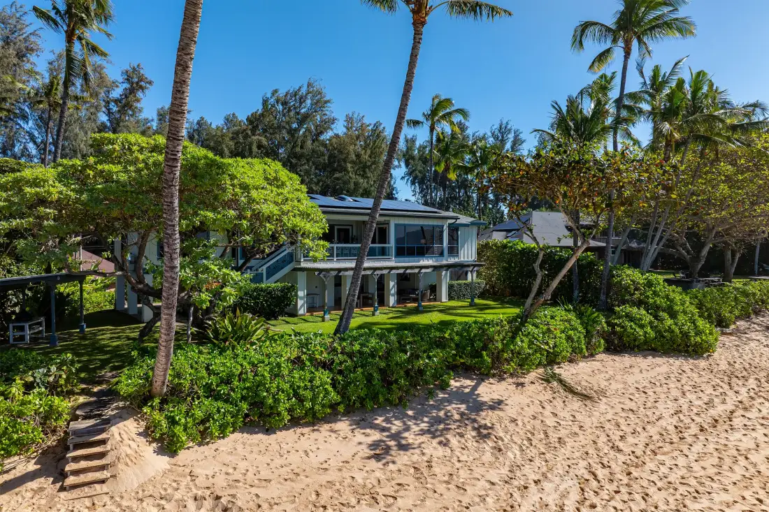 A modern building sits among tropical greenery beside a sandy beach.