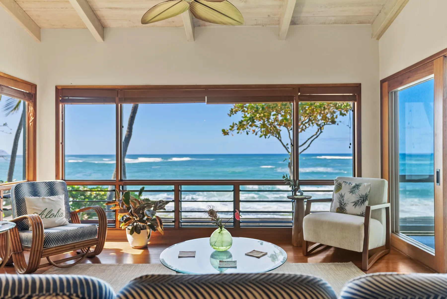A bright living room faces the ocean through wide windows, with seating around a glass table.
