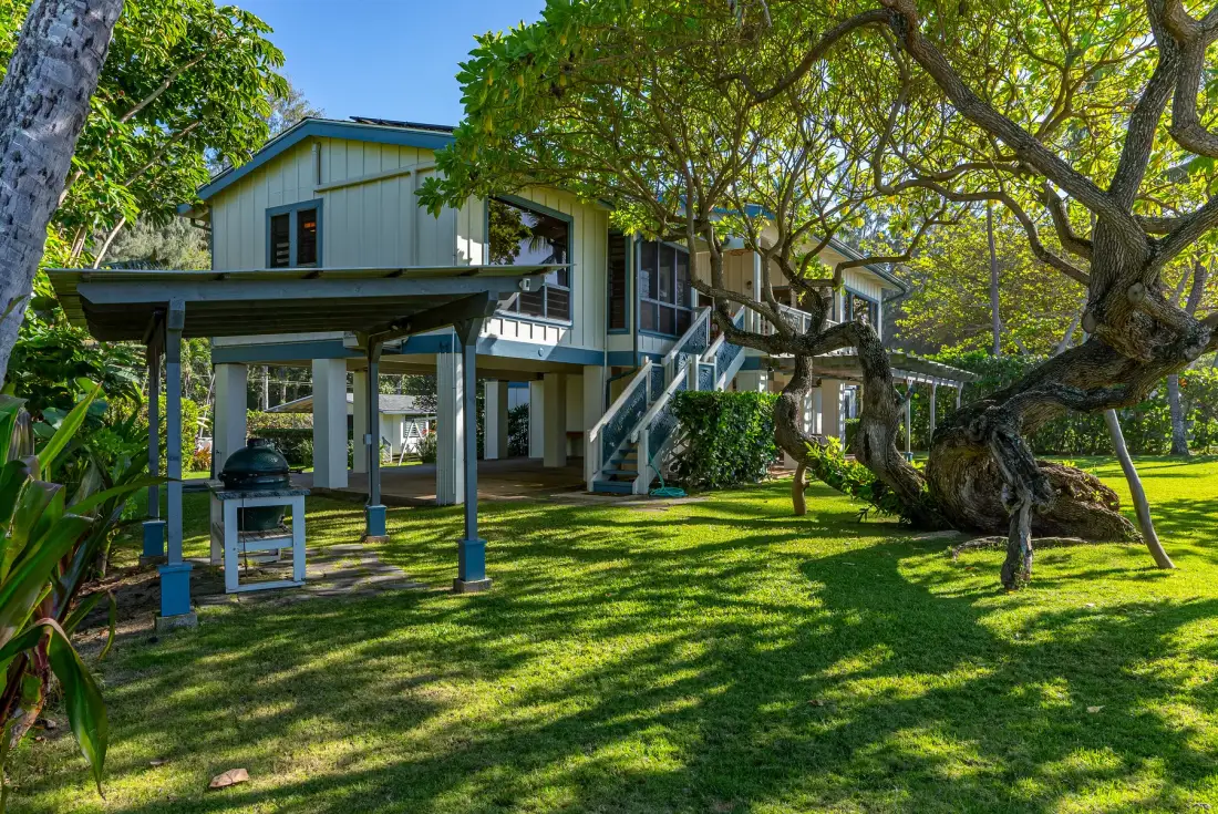 Casa elevada con una escalera exterior, bajo la sombra de árboles en un jardín con césped.