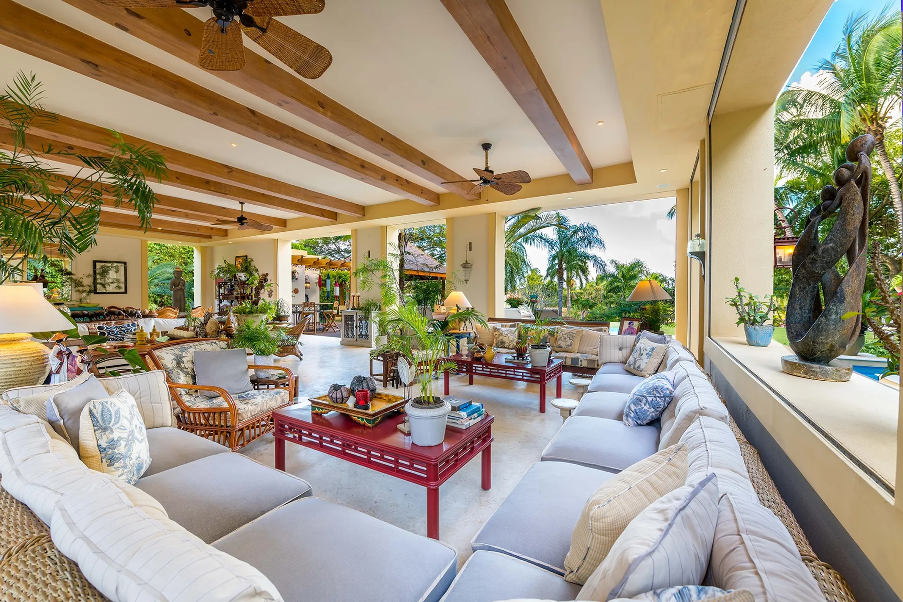 An open living room with light seating, red coffee tables, and views of tropical greenery.