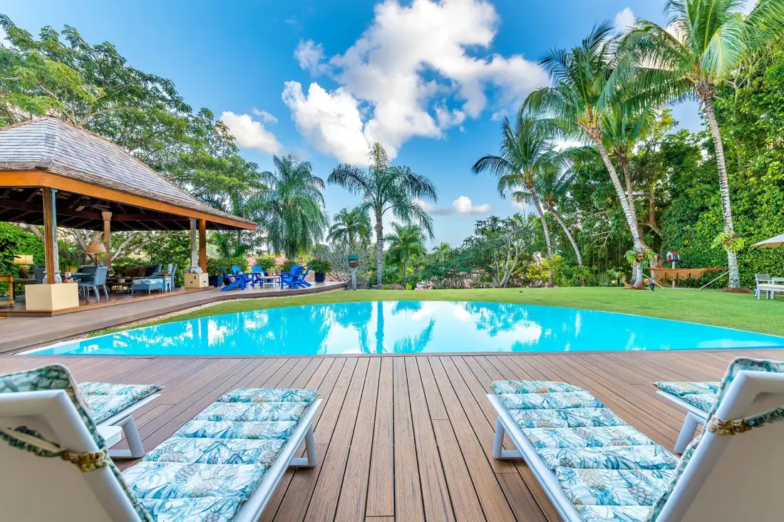 A tropical outdoor swimming pool is bordered by a wooden deck and palm trees.