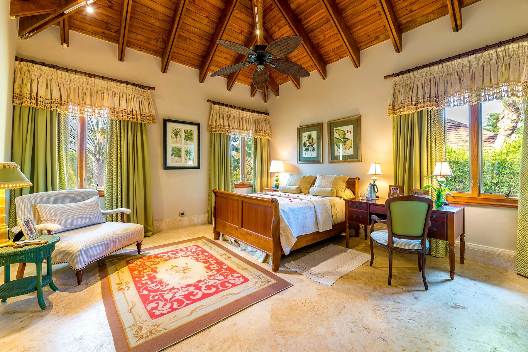 A bedroom with a large bed, seating, and windows overlooking greenery.