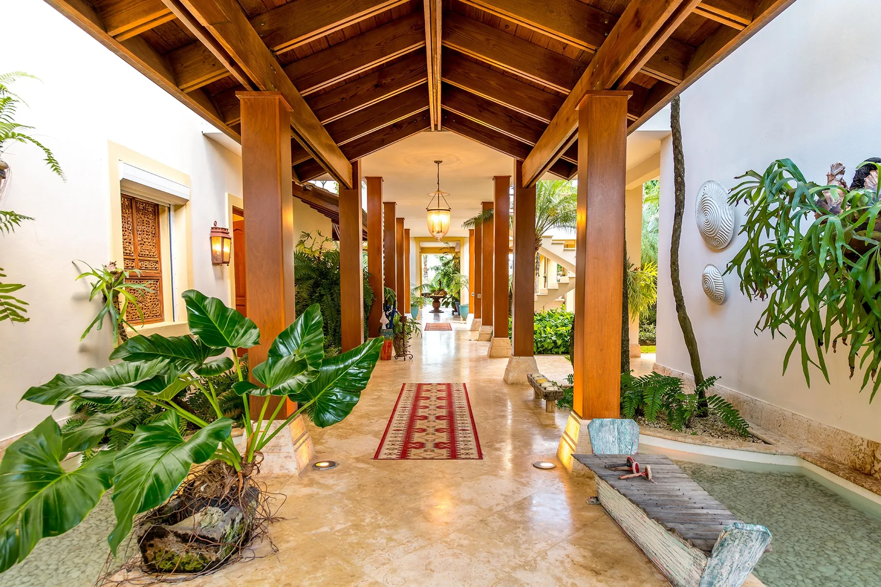 A covered walkway with wooden columns, hanging lights, and tropical plants.