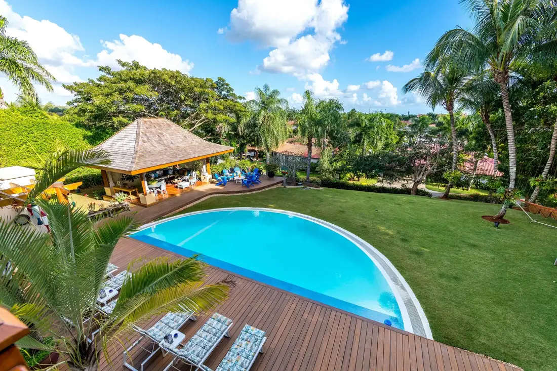 A tropical pool area with a wooden deck, lounge chairs, and a covered pavilion.