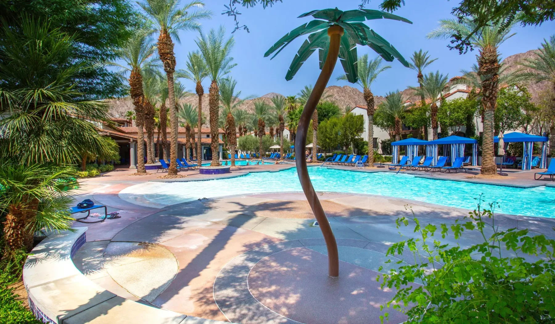 Una piscina exterior ajardinada está rodeada de palmeras y tumbonas azules.