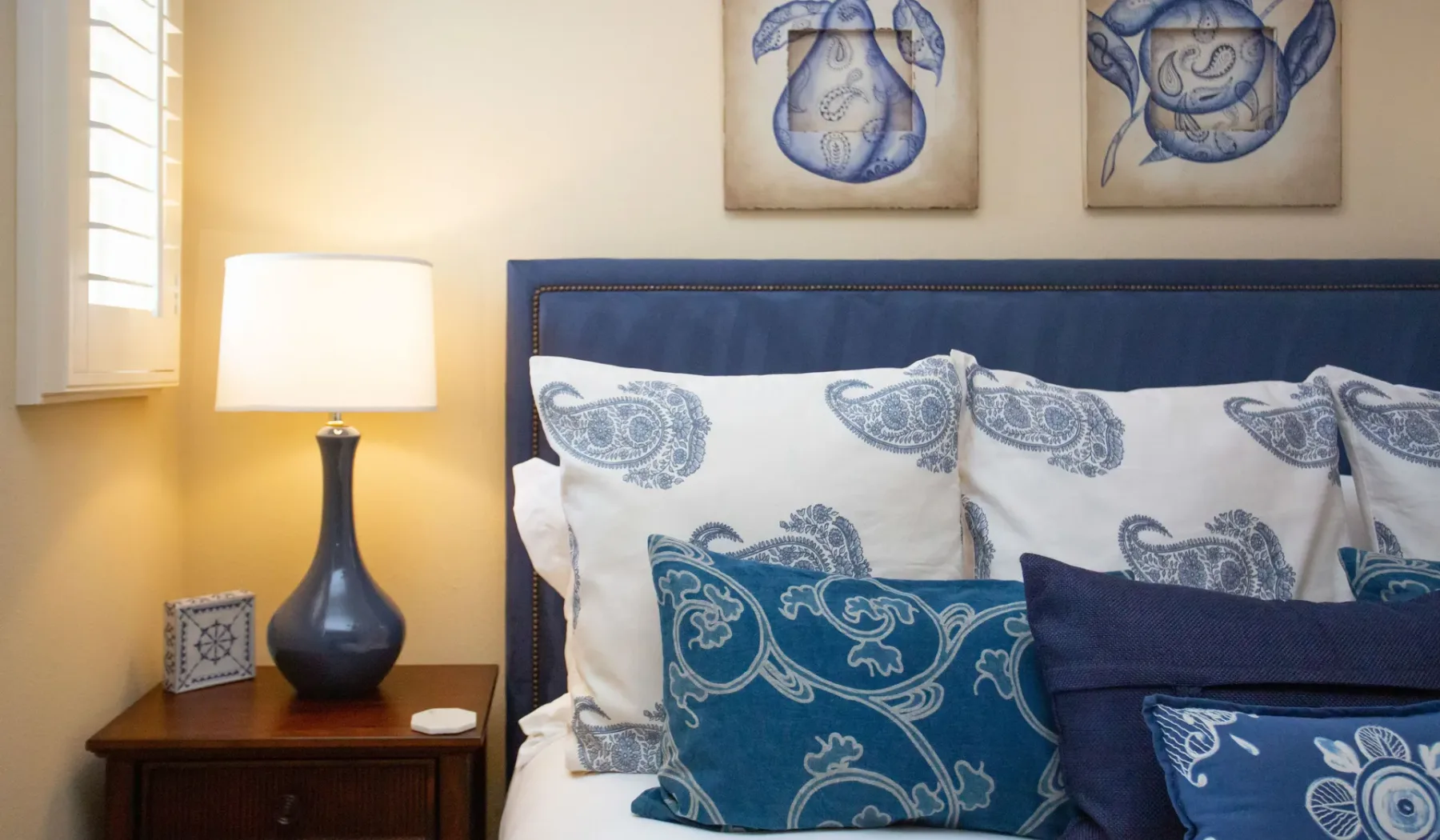 Un dormitorio con cabecero azul, almohadas estampadas y una lámpara de noche.
