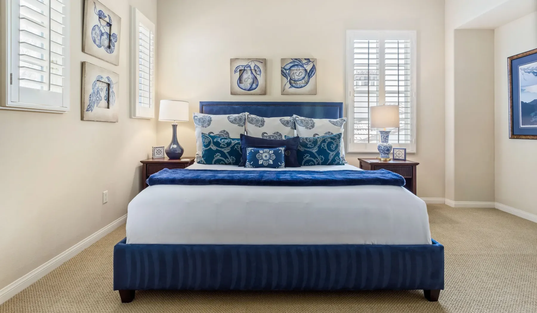 Un dormitorio con una cama azul, cojines estampados y ventanas con contraventanas.