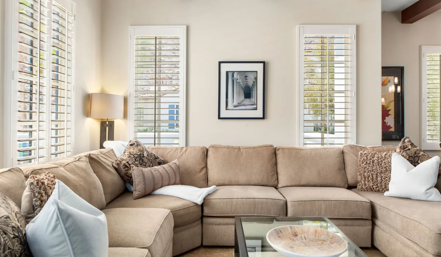 Una sala de estar con asientos beige, ventanas con contraventanas y una mesa de centro de vidrio.