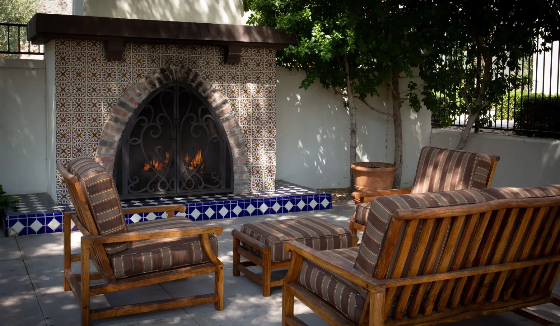 Una zona de estar al aire libre frente a una chimenea con azulejos, bajo árboles frondosos.