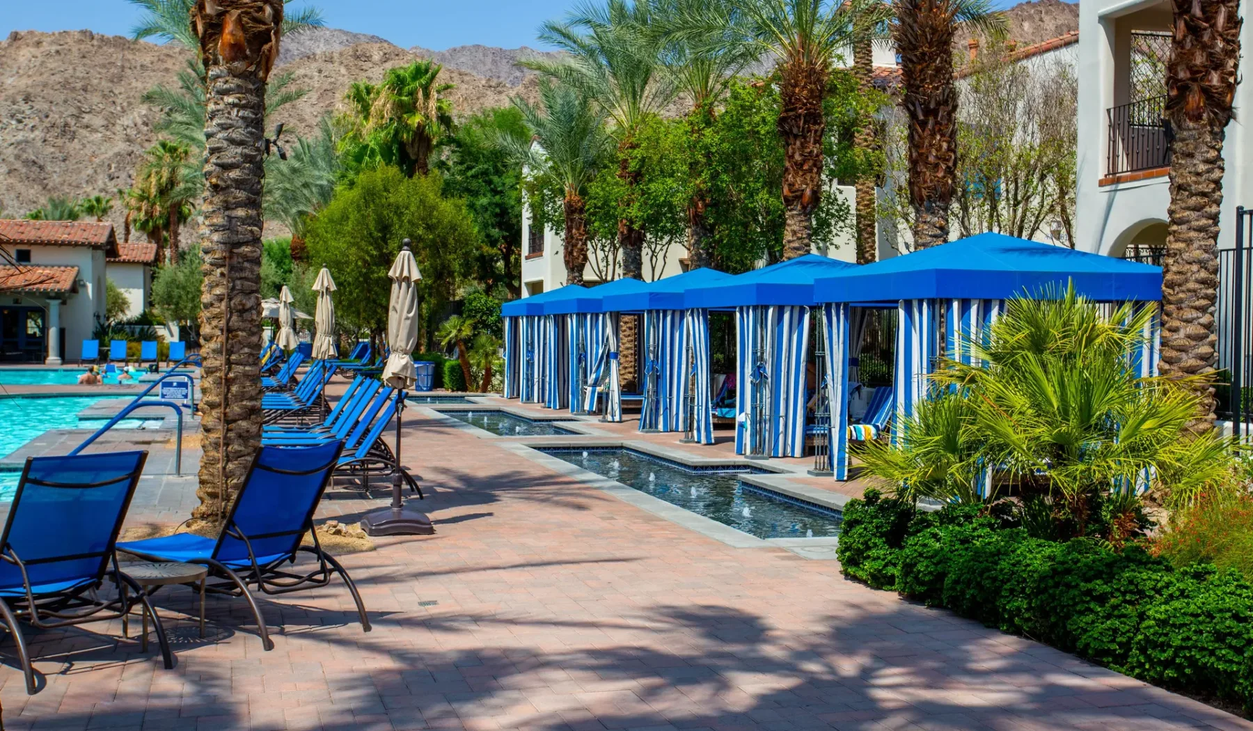 Una zona de piscina al aire libre tiene asientos azules, cabañas con sombra y palmeras.