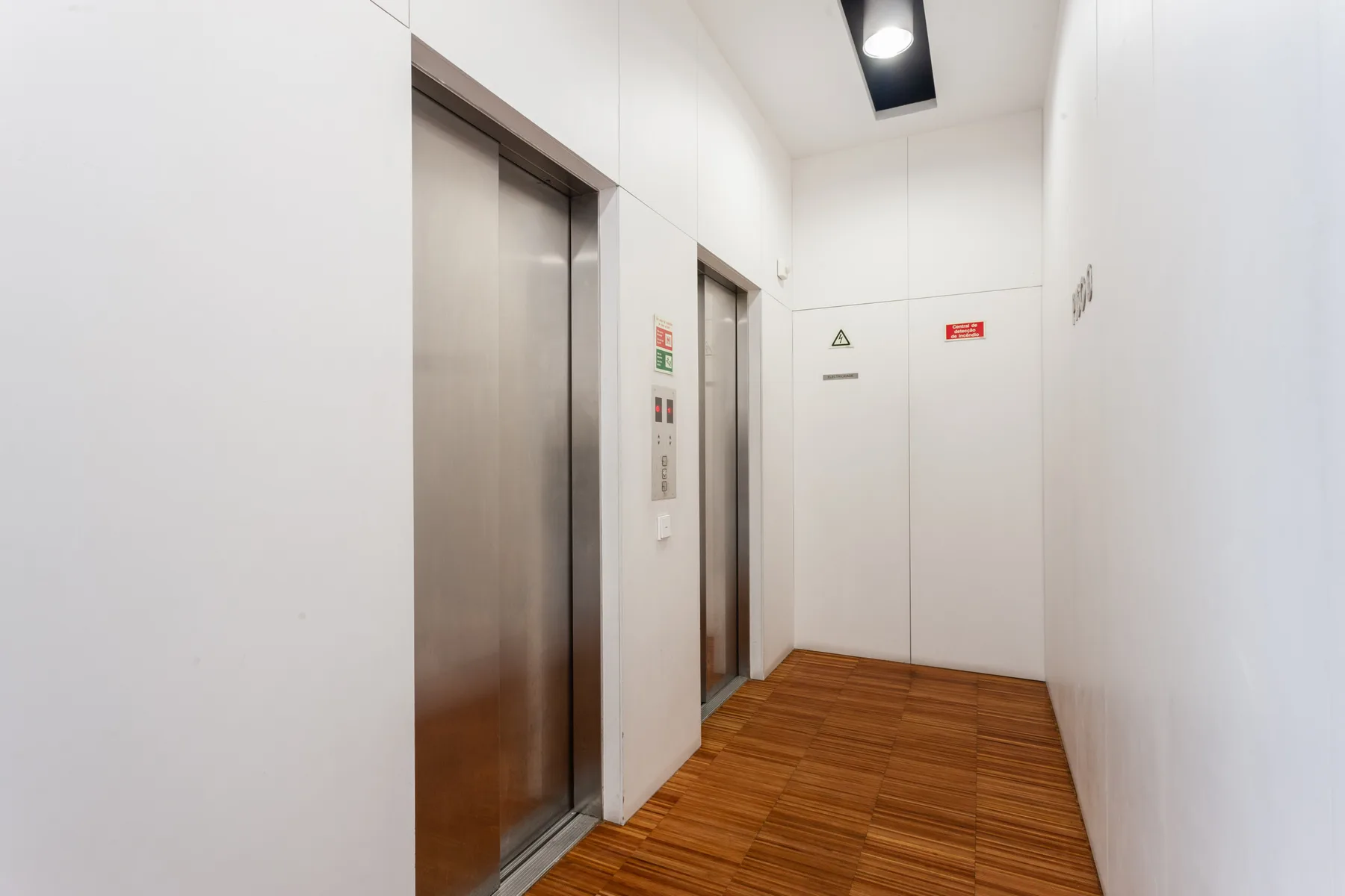 A bright elevator lobby with metallic elevator doors and a wood-patterned floor.
