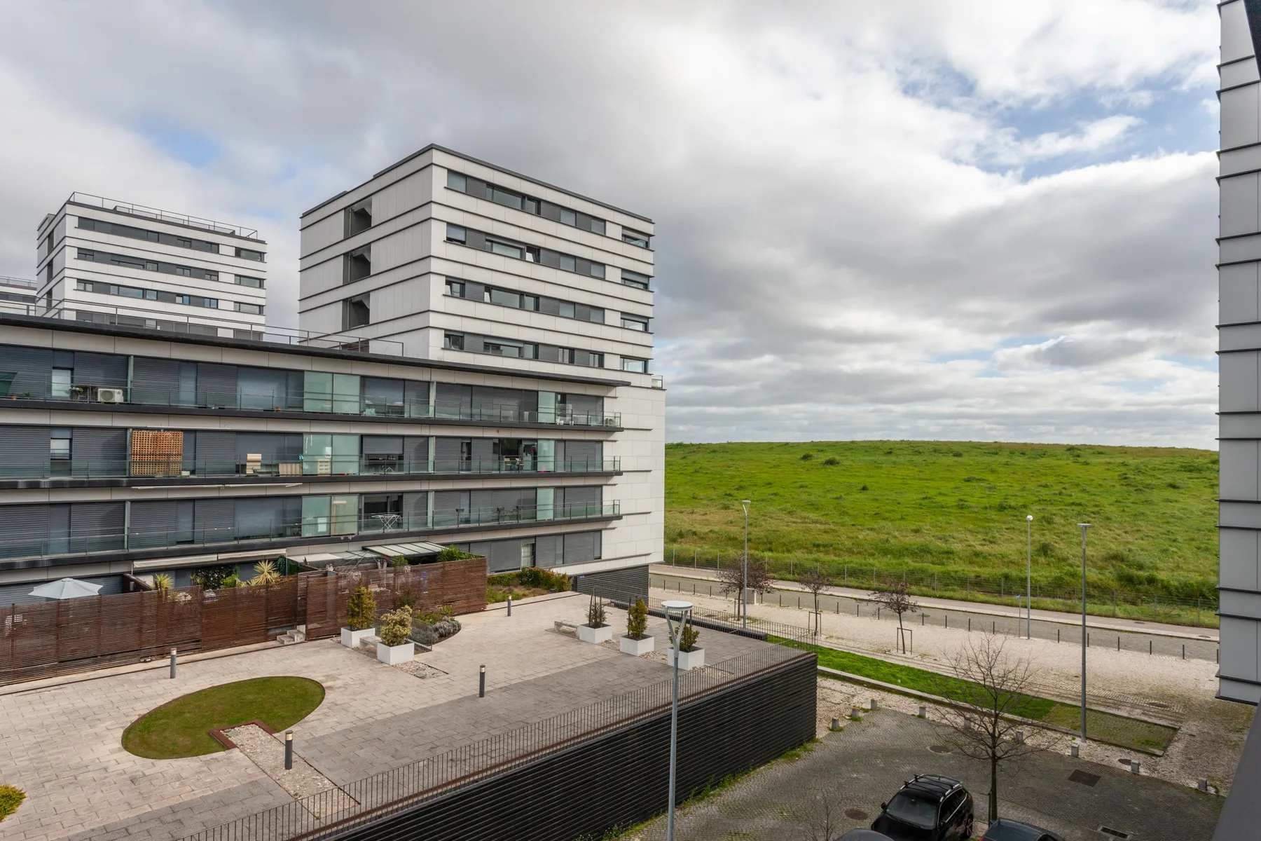 Modern apartment buildings overlook a broad green field and paved outdoor area.