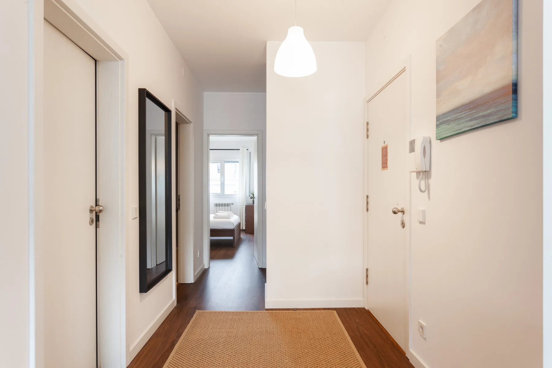 A bright apartment hallway with a woven rug and a bedroom visible at the end.