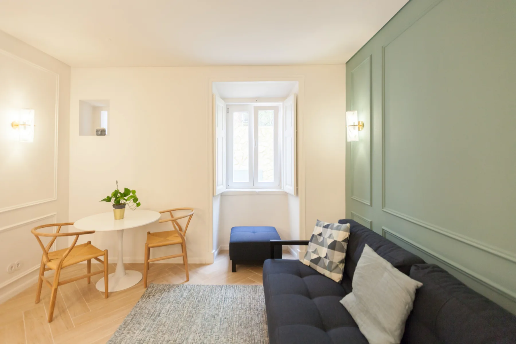 Una sala luminosa tiene un sofá oscuro, una mesa pequeña y una ventana.