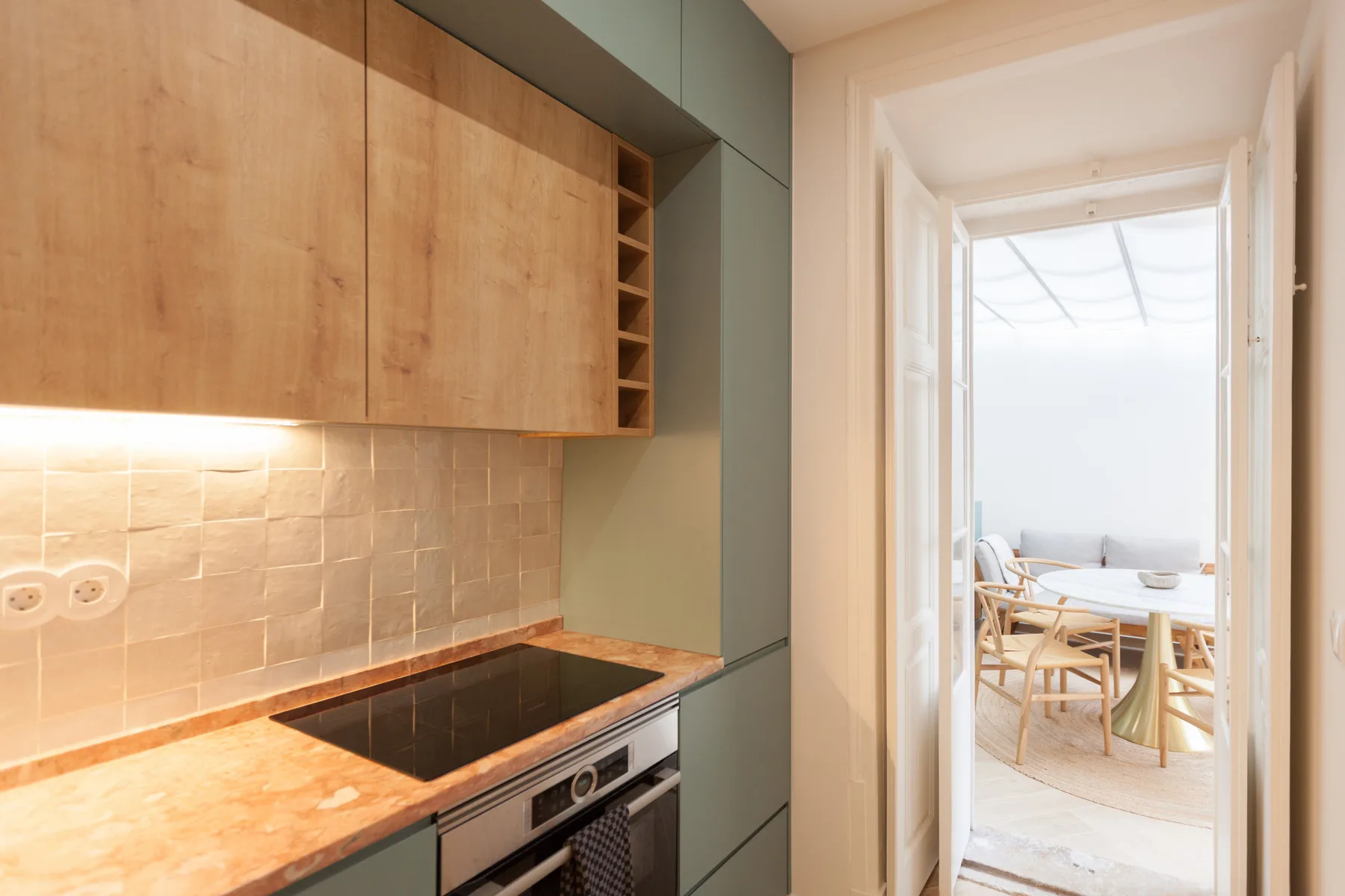 Una cocina compacta con armarios de madera y una placa negra da a un comedor luminoso.
