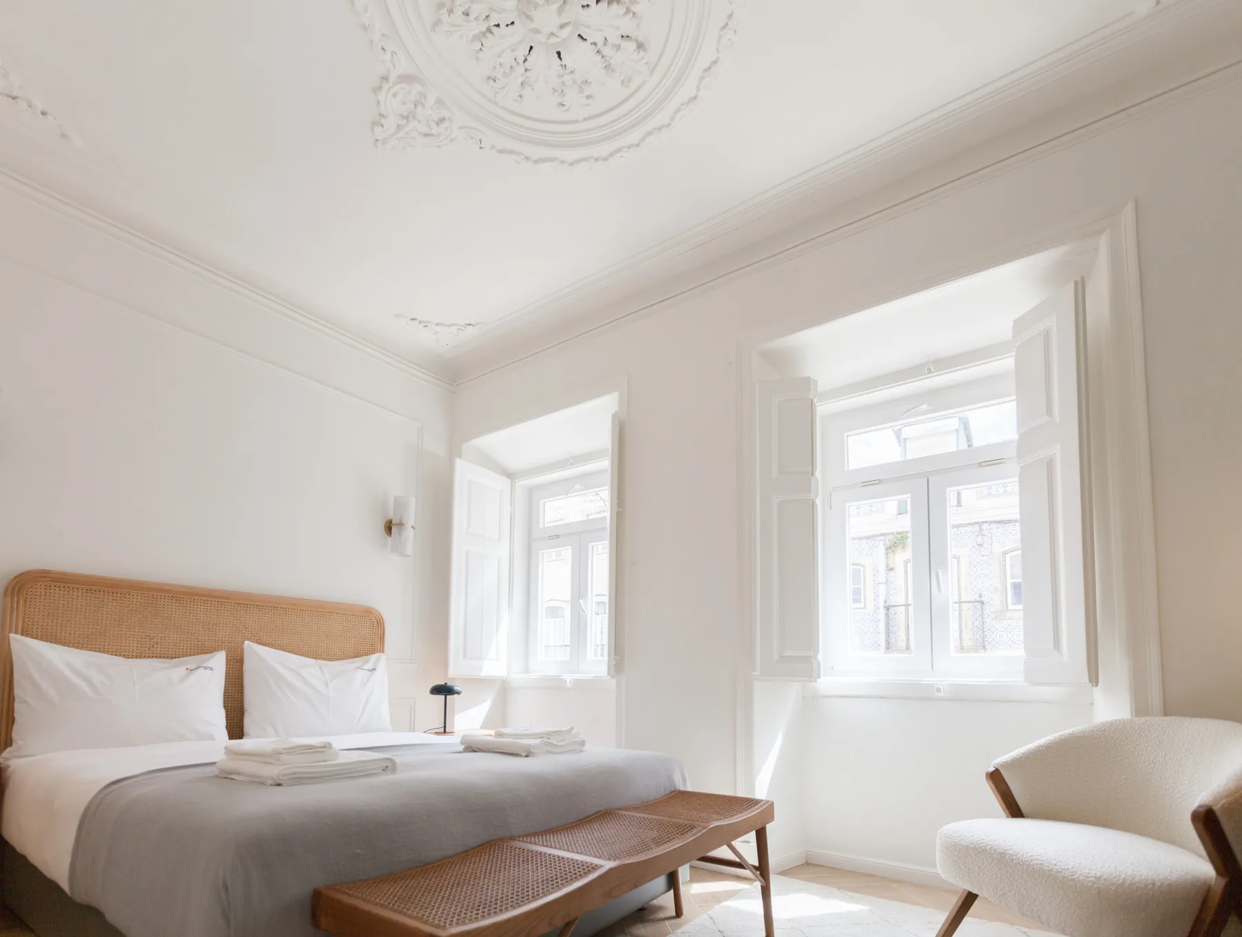 Un dormitorio luminoso con cama, banco de mimbre y ventanas grandes.