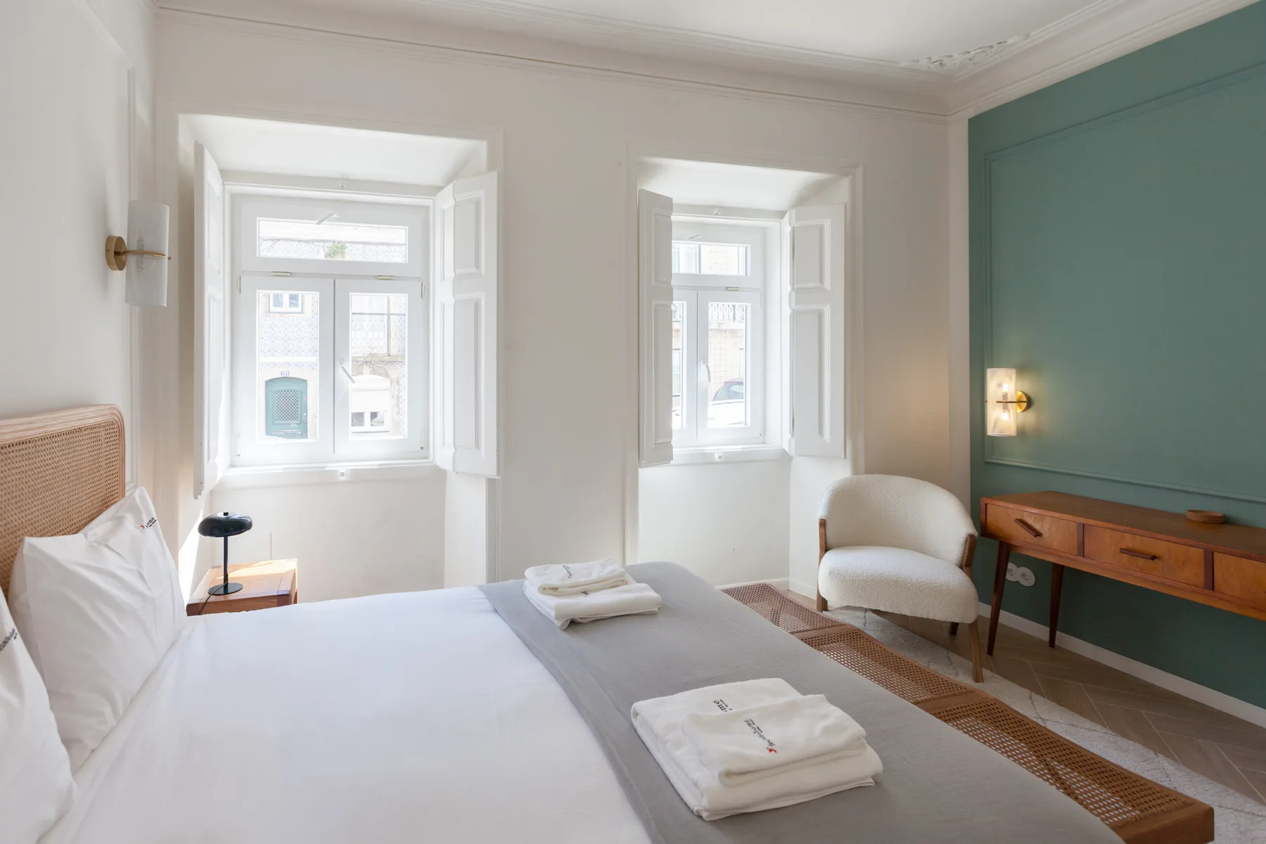 Dormitorio luminoso con una cama bien tendida, dos ventanas y una pequeña zona de estar.
