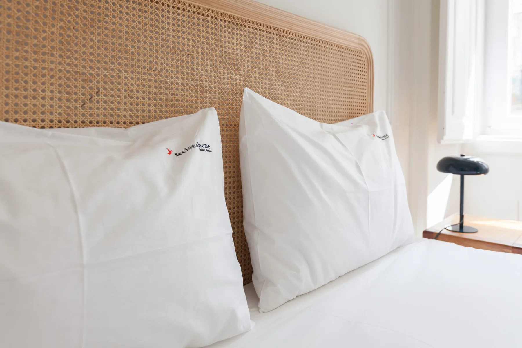 Una cama con almohadas blancas está junto a una ventana luminosa.
