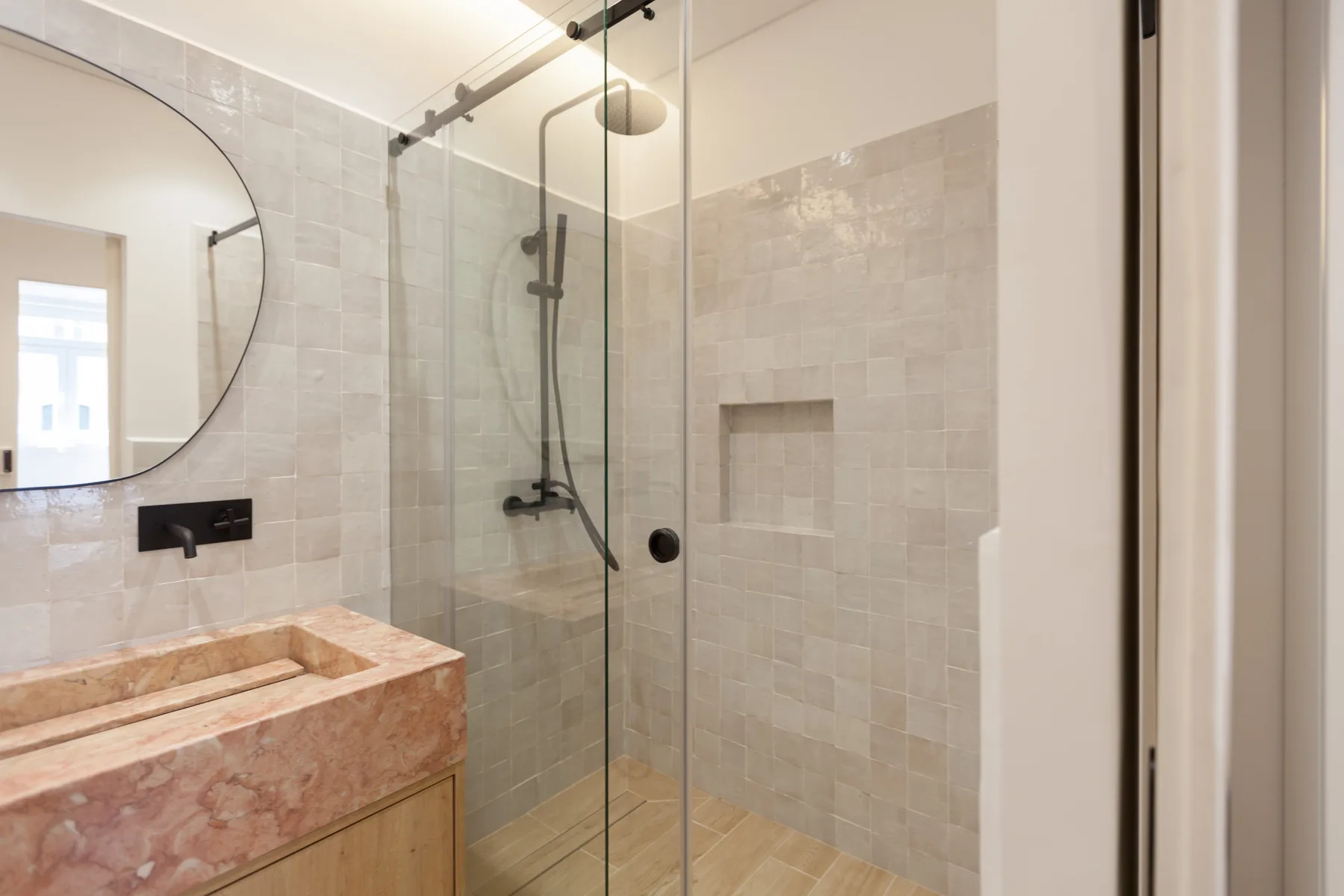 Un baño revestido de azulejos con ducha de vidrio, accesorios en la pared y un espejo redondo.
