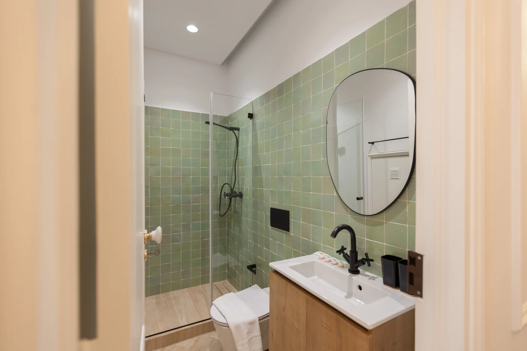 Un baño alicatado con ducha de vidrio y un lavabo bajo un espejo redondo.
