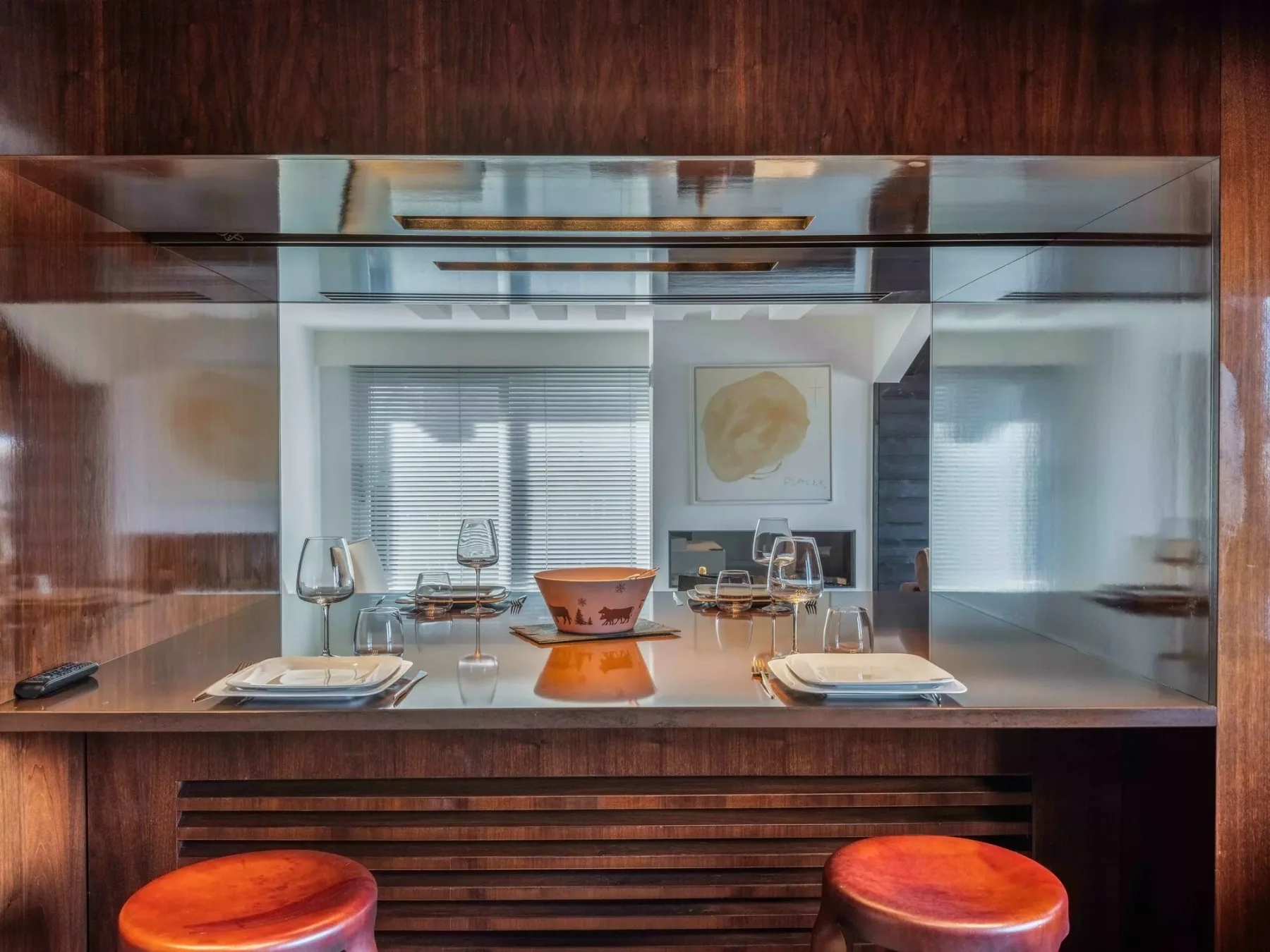 Una barra de comedor con puestos preparados y dos taburetes está frente a una sala luminosa con ventanas.