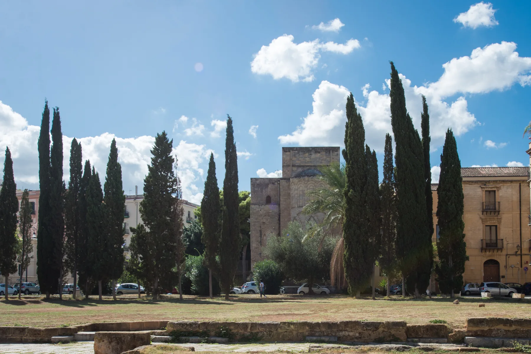 Una plaza soleada al aire libre con cipreses altos y edificios históricos de piedra.