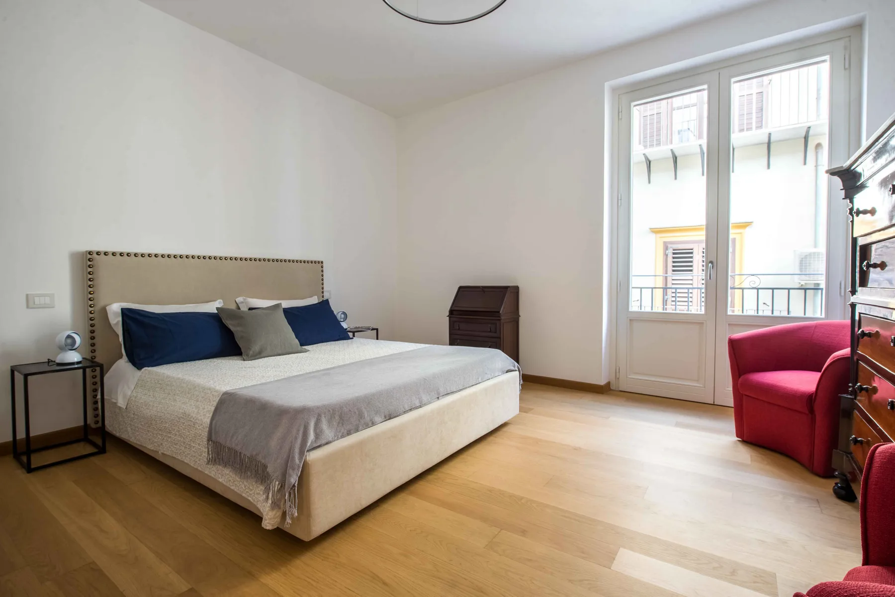 Un dormitorio luminoso con una cama tendida y una ventana con vistas a edificios cercanos.