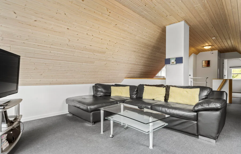Una sala de estar con asientos oscuros, una mesa de centro de vidrio y techo inclinado revestido de madera.