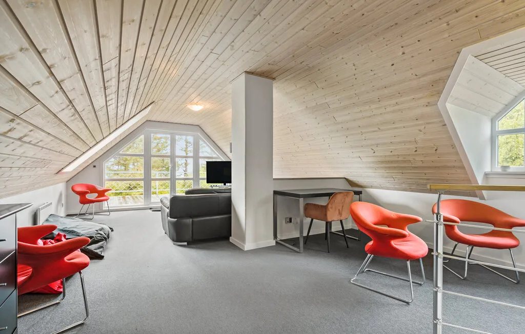 Una luminosa sala abuhardillada tiene asientos rojos, un sofá gris y grandes ventanas.